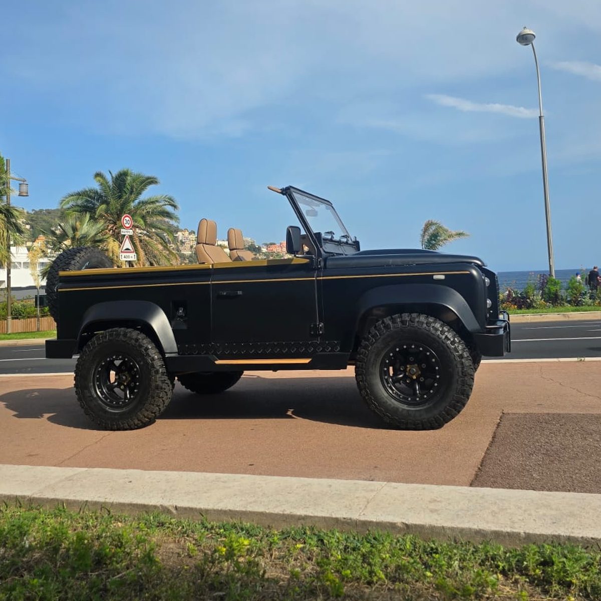 Land Rover Defender 90 V8 cabriolet noir mat restauré, vue de profil sur le bord de mer à Nice, ceinture de caisse bronze et jantes noires